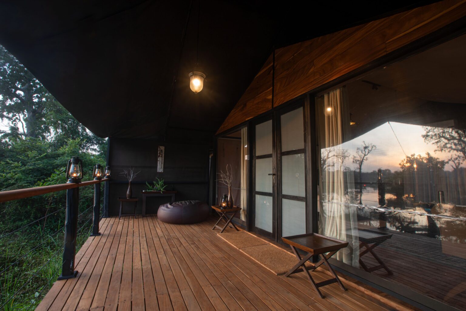 A timber deck along a tent above water, with the sun setting behind the trees